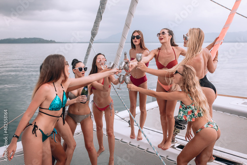 Happy slim girls in bright swimsuits and sunglasses clink glasses of champagne on a white yacht abroad on the open sea. Girly gatherings and a good time. The concept of an expensive vacation.