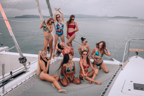 Posing on a yacht. Young tanned girlfriends in swimwear and sunglasses participate in a photo session. Someone is sitting, someone is standing. Holding glasses of champagne. Slim beauties on a cruise.