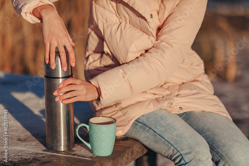 Cropped photograph of the outdoors. A girl in blue jeans and a pink down jacket opens an iron thermos for pouring tea. A cold winter day. The desire to warm up. The concept of keeping warm.