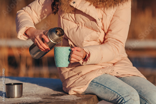 Close-up photo taken outdoors in winter. Women's hands hold an iron thermos and a blue cup to pour hot tea for warming. Background of the forest. A walk through the winter forest and a break for tea.