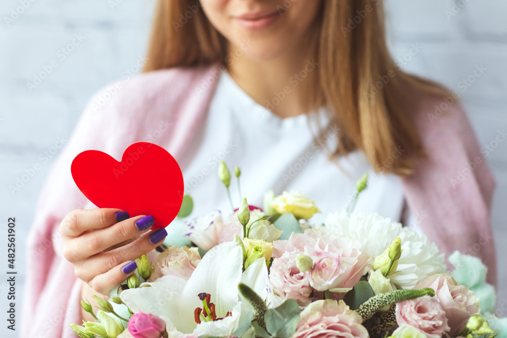 close-up of hands of smiling happy girl with card romantic letter in shape of red heart and large bouquet of flowers, congratulations on Valentine's day or women's Day. Surprised. Delivery presents
