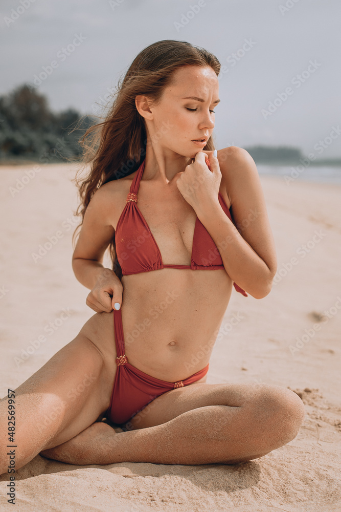 Exotic beach photo shoot. A young adorable woman poses sitting on the sand in a red bikini with her eyes closed. Blurry background of the sky, ocean and trees. Summer landscape. Exotic beach photo shoot. A young adorable woman poses sitting on the sand in a red bikini with her eyes closed. Blurry background of the sky, ocean and trees. Summer landscape.