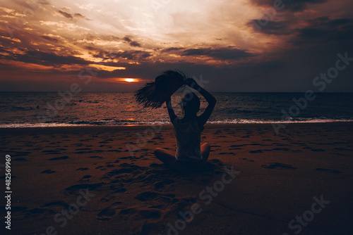 Photo shoot at sunset. A young stunning woman is sitting with her back on the sand and posing with a straw hat in her raised hands. An atmospheric tropical landscape with the setting sun.