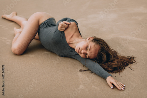 Photo shoot on the beach sand. A very beautiful young and sexy woman in a stylish gray bodysuit lies on the hot sand and poses arching with her eyes closed. The concept of the beauty. photo