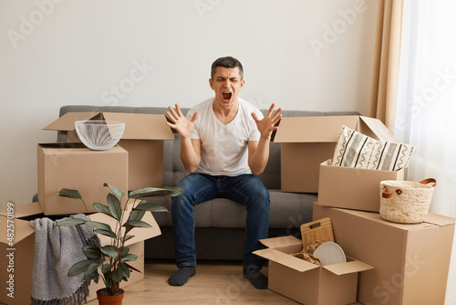 Portrait of angry man wearing white T-shirt and jeans sitting on sofa surrounded with cardboard boxes, raised arms, screaming with aggressive, moving and relocating.