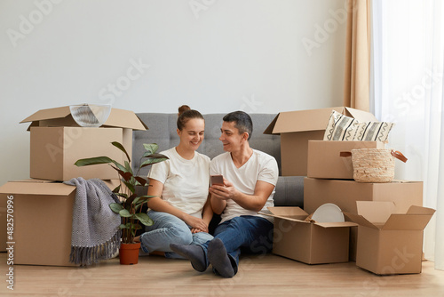 Indoor shot of young couple in love moving in a new apartment, sitting on the floor near, planning to redecorate their new home, man holding smart phone, showing ideas to his wife.