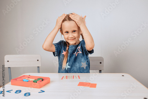 Portrait of a cute, affectionate and kind girl sitting at a table surrounded by puzzles and putting her hands on her head. Shocked girl. Emotion concept. photo with noise