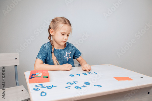 Portrait of a focused intelligent cool child sitting at a white table with numbers in the correct order and a girl pointing to the numbers, memorizing them. Development process. Study concept. 