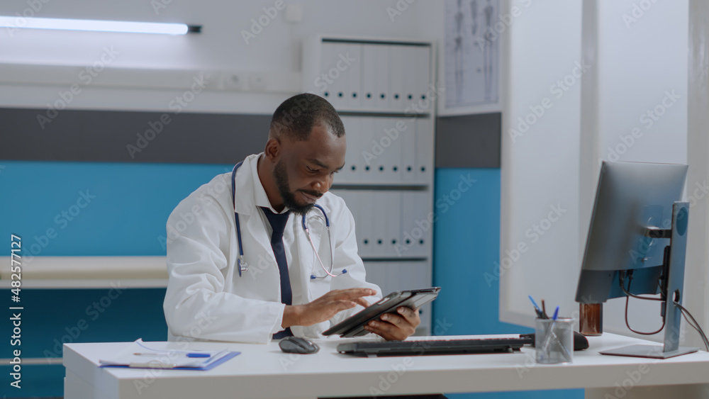 African american therapist doctor sitting at desk in hospital office holding tablet computer analyzing medical documents typing sickness expertise. Physician man developing healthcare treatment