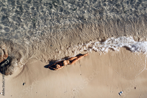 Overhead view of a sexy slim and attractive model in a black bikini lying on the wet sand of the beach and the waves of the sea hitting her tanned body. Perfect vacation concept