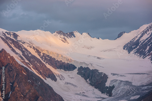 Dramatic aerial view to high snow mountain range in early morning at dawn. Awesome scenery with sunlit snow mountains in cloudy sky at sunrise. Scenic landscape with large glacier in sunrise colors.