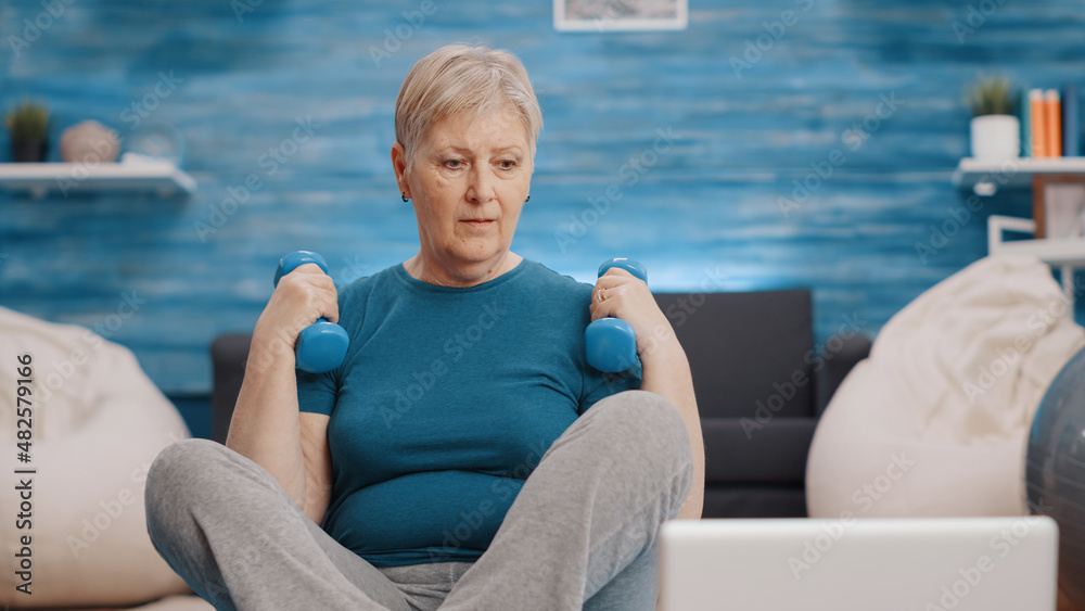 Fototapeta premium Close up of pensioner training with dumbbells on yoga mat, looking at laptop with workout video. Retired woman using weights lifting to exercise and following online workout on computer