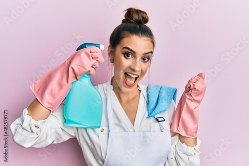 Beautiful brunette young woman wearing apron holding cleaning spray pointing to head screaming proud  celebrating victory and success very excited with raised arms
