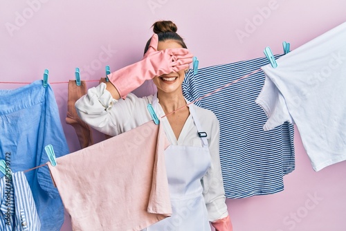 Beautiful brunette young woman washing clothes at clothesline smiling and laughing with hand on face covering eyes for surprise. blind concept.