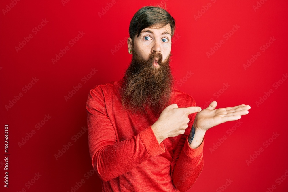 Redhead man with long beard pointing open palm with finger making fish face with mouth and squinting eyes, crazy and comical.