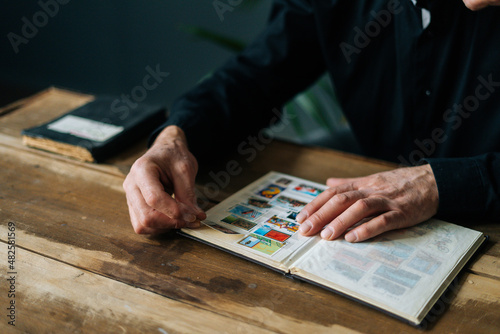 Close-up of unrecognizable senior adult man inserting postage stamps at collection book sitting at wooden table, selective focus. Mature aged man looking album with old stamps philately at home. photo