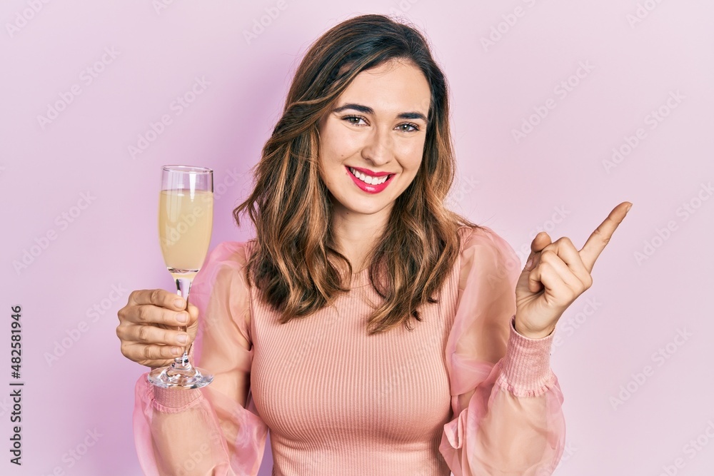 Young hispanic girl drinking a glass of sparkling champagne smiling happy pointing with hand and finger to the side Young hispanic girl drinking a glass of sparkling champagne smiling happy pointing with hand and finger to the side