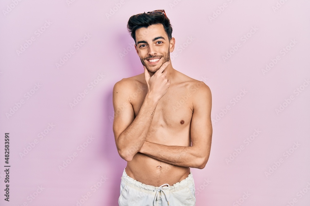 Fototapeta premium Young hispanic man wearing swimwear shirtless looking confident at the camera smiling with crossed arms and hand raised on chin. thinking positive.
