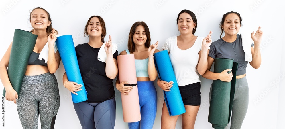 Obraz premium Group of women holding yoga mat standing over isolated background gesturing finger crossed smiling with hope and eyes closed. luck and superstitious concept.