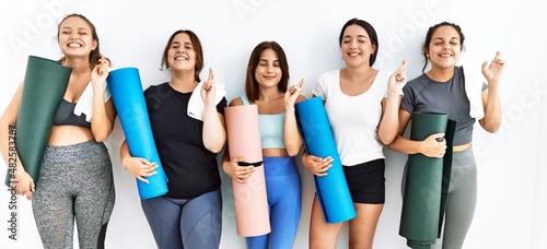 Group of women holding yoga mat standing over isolated background gesturing finger crossed smiling with hope and eyes closed. luck and superstitious concept.