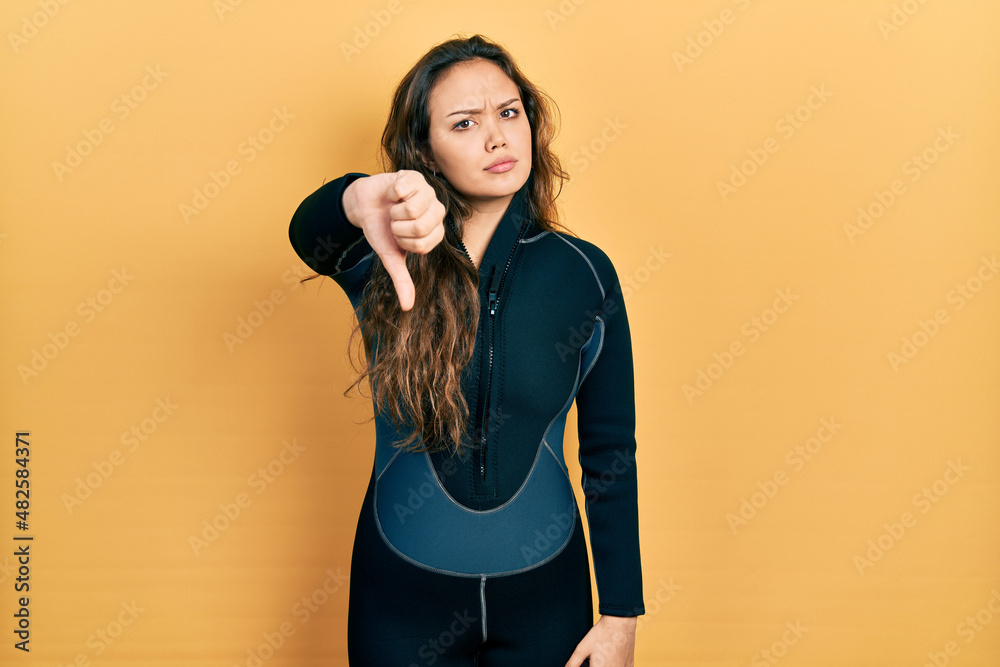 Young hispanic girl wearing diver neoprene uniform looking unhappy and angry showing rejection and negative with thumbs down gesture. bad expression.