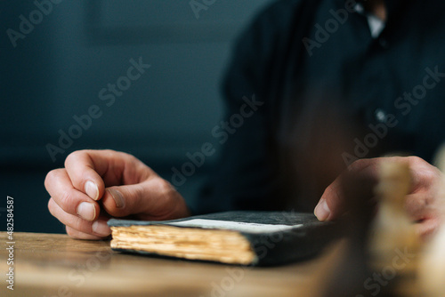 Close-up cropped shot of unrecognizable senior aged male sitting at wooden table with old book, selective focus. Closeup of mature man reading older notebook with notes, nostalgic about past life.