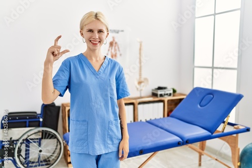 Beautiful caucasian physiotherapist woman working at pain recovery clinic smiling and confident gesturing with hand doing small size sign with fingers looking and the camera. measure concept.
