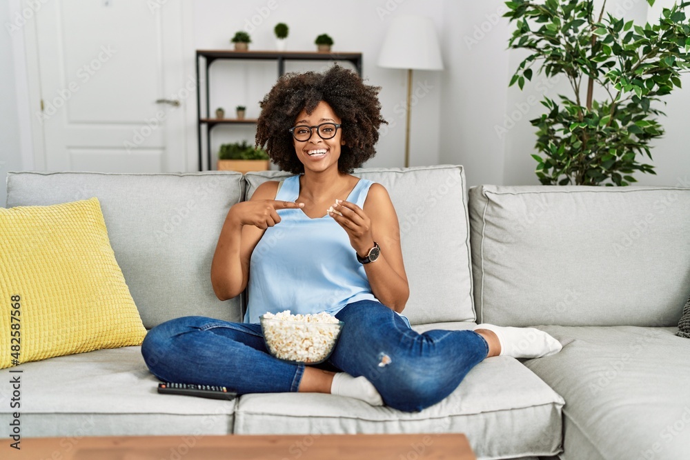 African american woman with afro hair sitting on the sofa eating popcorn at home smiling happy pointing with hand and finger African american woman with afro hair sitting on the sofa eating popcorn at home smiling happy pointing with hand and finger