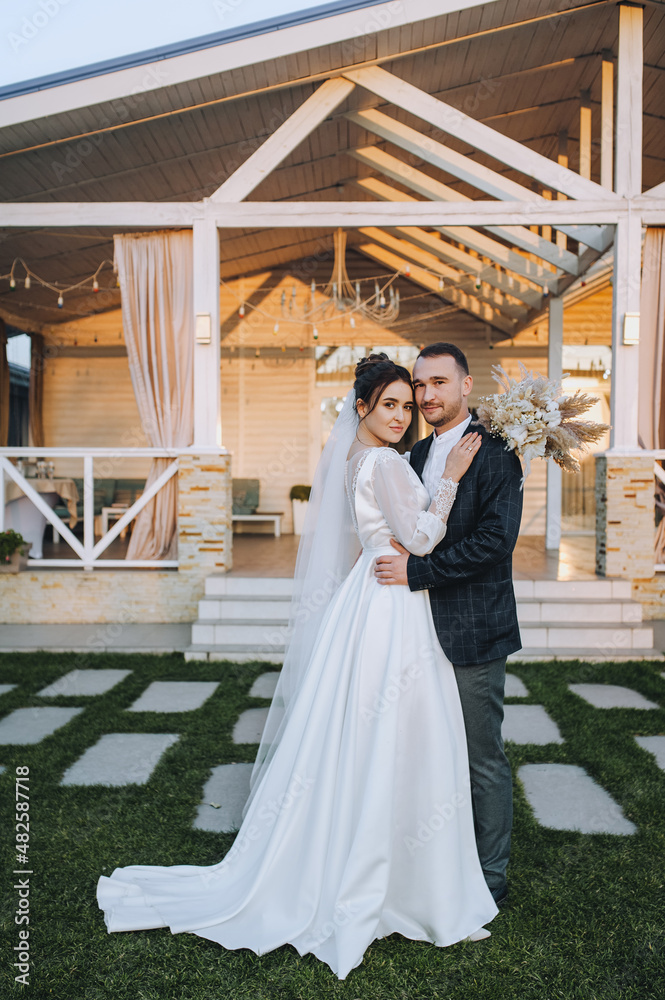 A young groom and a beautiful, smiling brunette bride in a white dress are standing embracing on the green grass, in nature, against the backdrop of a cottage, at home. Wedding portrait, photography. A young groom and a beautiful, smiling brunette bride in a white dress are standing embracing on the green grass, in nature, against the backdrop of a cottage, at home. Wedding portrait, photography.