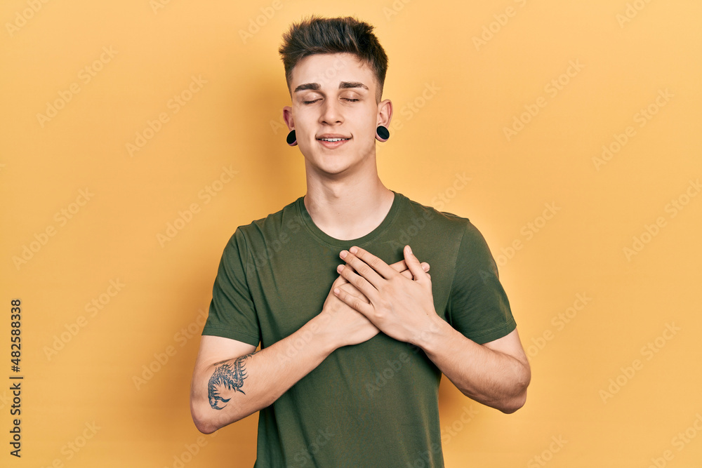 Young caucasian boy with ears dilation wearing casual green t shirt smiling with hands on chest with closed eyes and grateful gesture on face. health concept. Young caucasian boy with ears dilation wearing casual green t shirt smiling with hands on chest with closed eyes and grateful gesture on face. health concept.