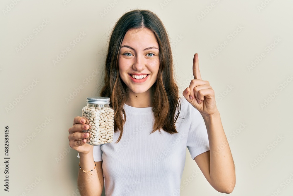 Young brunette woman holding bottle with raw white beans smiling with an idea or question pointing finger with happy face, number one Young brunette woman holding bottle with raw white beans smiling with an idea or question pointing finger with happy face, number one