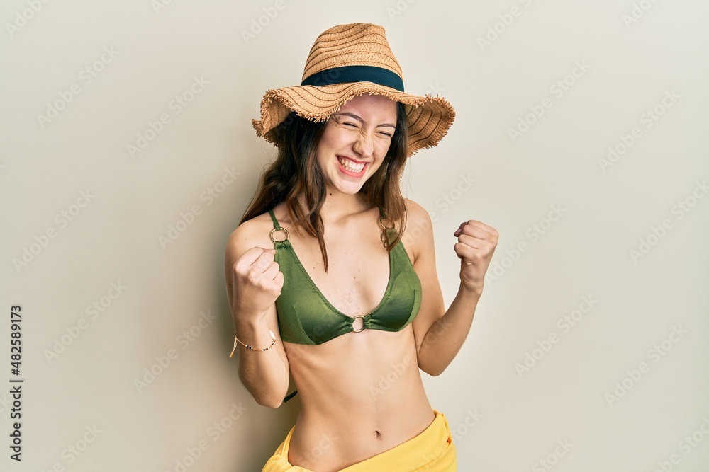 Young brunette woman wearing bikini excited for success with arms raised and eyes closed celebrating victory smiling. winner concept. Young brunette woman wearing bikini excited for success with arms raised and eyes closed celebrating victory smiling. winner concept.