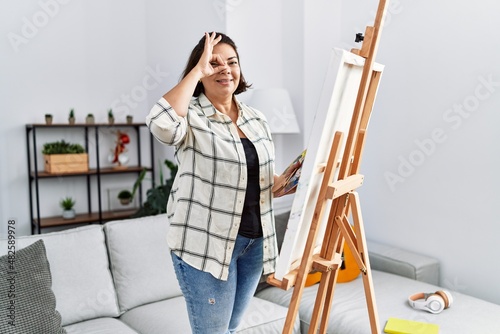 Middle age hispanic woman drawing by painter easel stand at home smiling happy doing ok sign with hand on eye looking through fingers