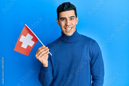 Handsome hispanic man holding switzerland flag looking positive and happy standing and smiling with a confident smile showing teeth