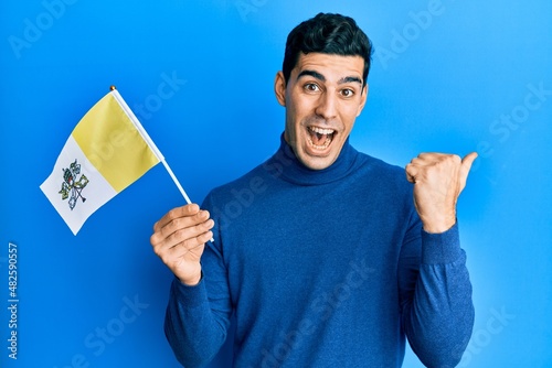 Handsome hispanic man holding vatican city flag pointing thumb up to the side smiling happy with open mouth