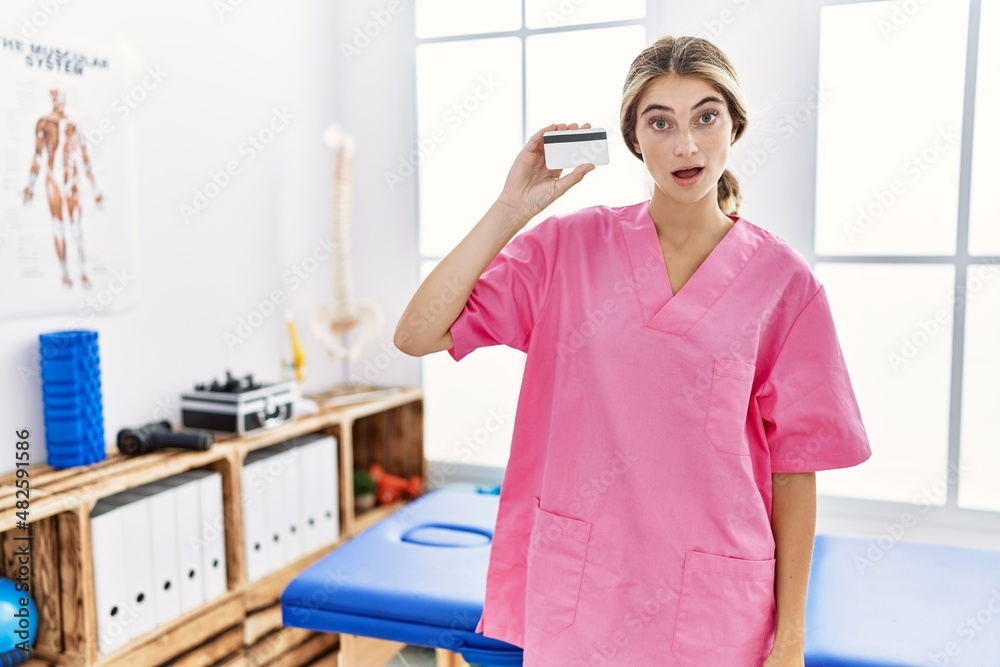 Young physiotherapist woman working at pain recovery clinic holding credit card scared and amazed with open mouth for surprise, disbelief face