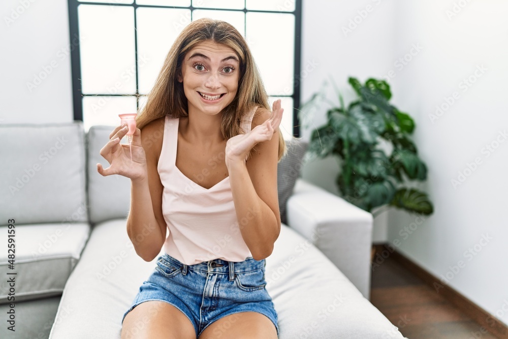 Young blonde woman holding menstrual cup celebrating victory with happy smile and winner expression with raised hands