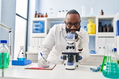 Young african american man wearing scientist uniform using microscope at laboratory