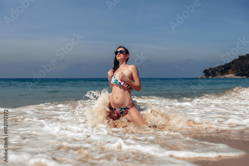 Lovely adorable woman with a perfect body in a bikini and glasses is kneeling on the beach near the sea and the waves are beating on her body. Deep sea concept.