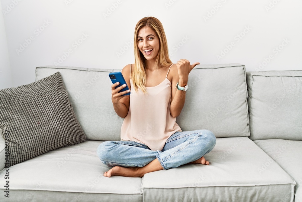 Blonde beautiful young woman sitting on the sofa at home using smartphone pointing to the back behind with hand and thumbs up, smiling confident Blonde beautiful young woman sitting on the sofa at home using smartphone pointing to the back behind with hand and thumbs up, smiling confident