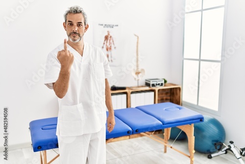 Middle age hispanic therapist man working at pain recovery clinic showing middle finger  impolite and rude fuck off expression