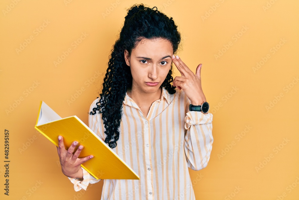Young hispanic woman with curly hair holding book shooting and killing oneself pointing hand and fingers to head like gun, suicide gesture. Young hispanic woman with curly hair holding book shooting and killing oneself pointing hand and fingers to head like gun, suicide gesture.