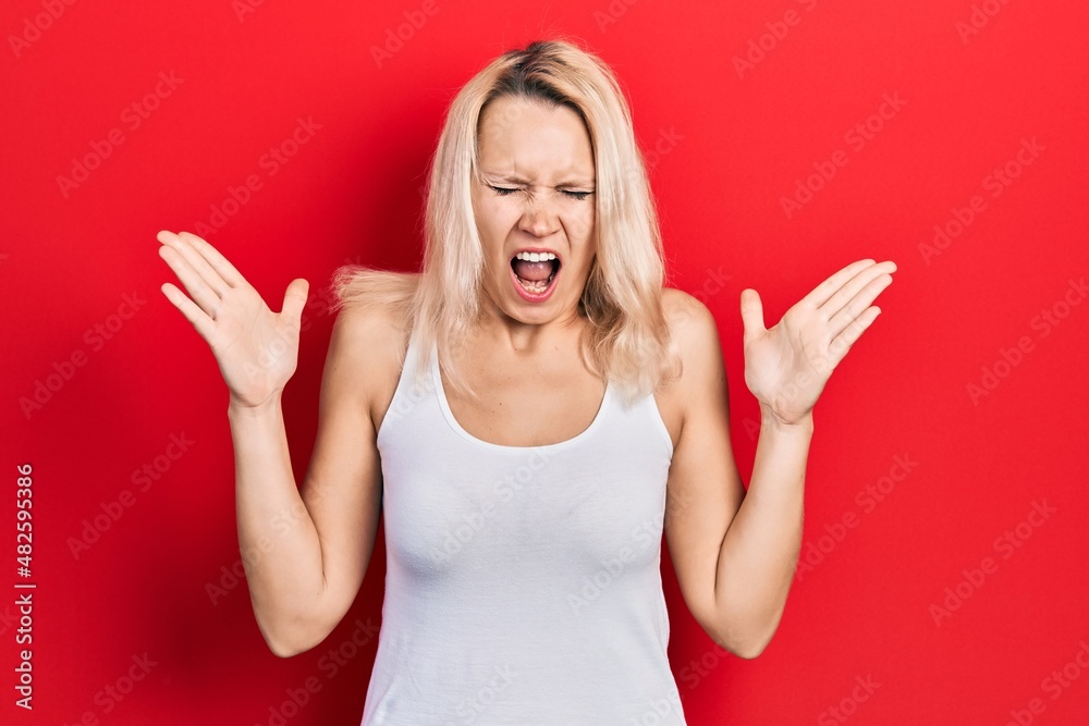 Beautiful caucasian blonde woman wearing casual white t shirt celebrating mad and crazy for success with arms raised and closed eyes screaming excited. winner concept Beautiful caucasian blonde woman wearing casual white t shirt celebrating mad and crazy for success with arms raised and closed eyes screaming excited. winner concept