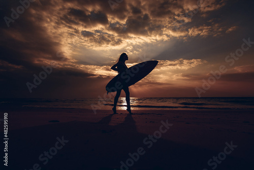 Silhouette of the surfer slender female model with surfboard in her hand on the sandy beach at sunset. Surfer and ocean. Side view.