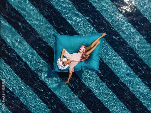 Top view of an adorable skinny girl in a bathrobe and with a towel on her head and sunglasses, floating in a pool on an inflatable mattress. Overflowing water. Summertime concept
