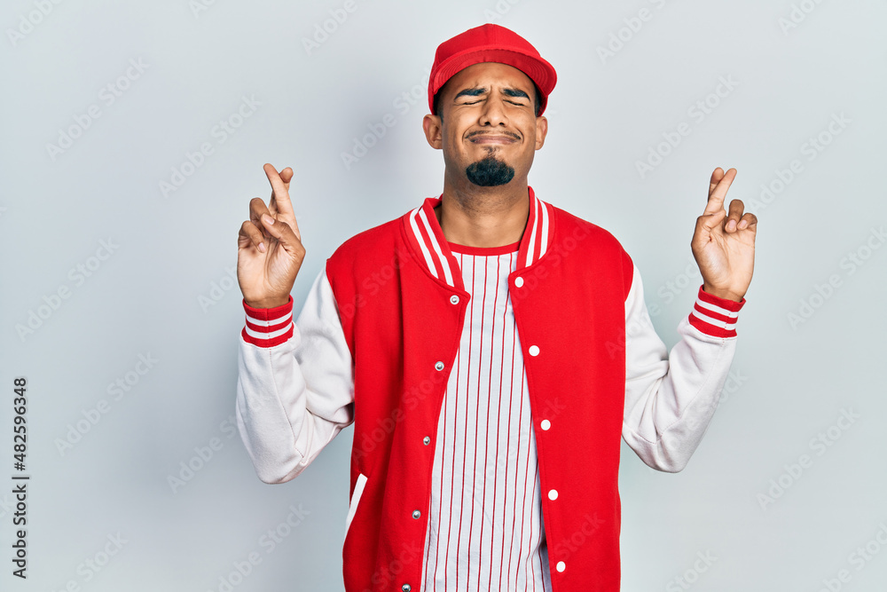 Young african american man wearing baseball uniform gesturing finger crossed smiling with hope and eyes closed. luck and superstitious concept.