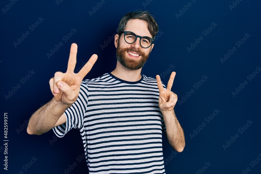 Caucasian man with beard wearing striped t shirt and glasses smiling looking to the camera showing fingers doing victory sign. number two.