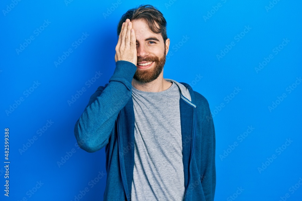 Caucasian man with beard wearing casual sweatshirt covering one eye with hand, confident smile on face and surprise emotion. Caucasian man with beard wearing casual sweatshirt covering one eye with hand, confident smile on face and surprise emotion.