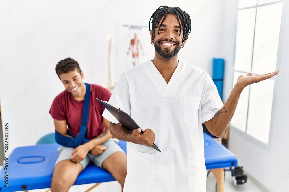 Obraz premium Young hispanic man working at pain recovery clinic with a man with broken arm smiling cheerful presenting and pointing with palm of hand looking at the camera.