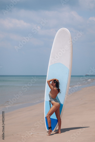 Vertical photo of a slim, lithe lady with mouth-watering curves, wearing a form-fitting bikini and posing next to a huge surfboard. Sea in the background. Summer concept